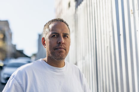 Kris Taute stands outdoors in a white t-shirt, looking sideways with a serious expression