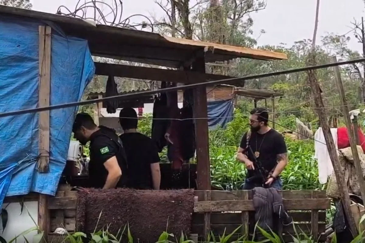 Foto colorida de movimentação de policiais em frente à barraco de madeira palco de homicídio.