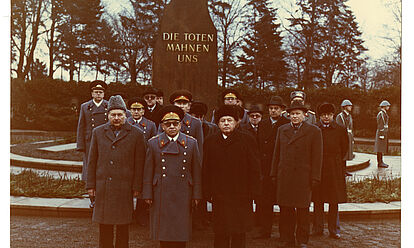 Eine Abordnung von Offizieren der Stasi und des sowjetischen KGB in Uniform und Zivil vor einer Stele auf der steht 'Die Toten mahnen uns'