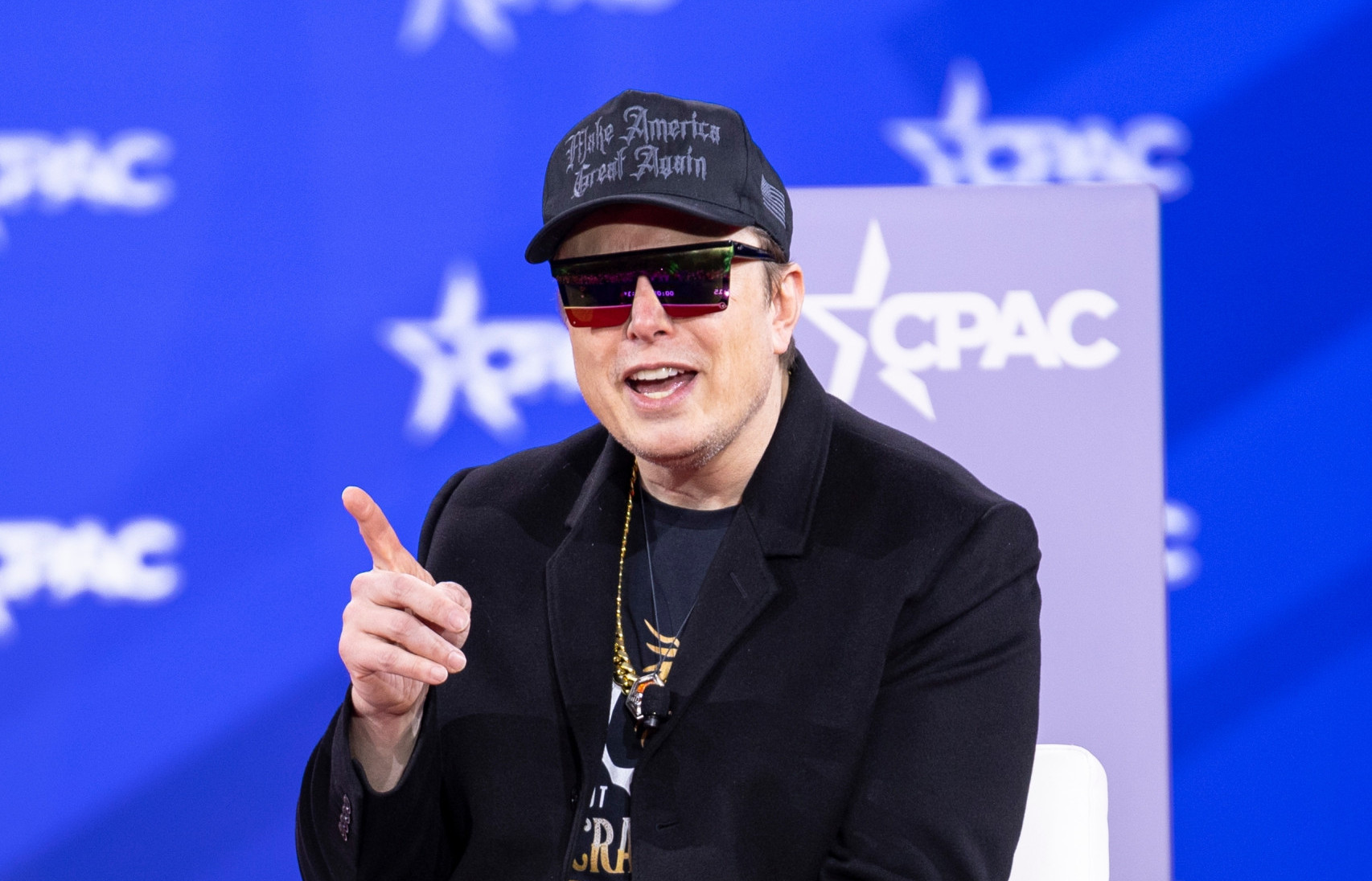 Elon Musk sentado, de óculos escuros, boné, falando e com o dedo indicador direito em riste