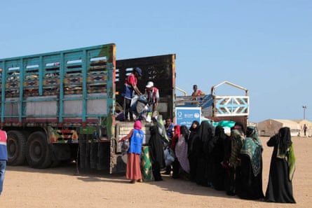 A view of a large truck in a desert with tents in the background where worker are handing supplies down to a queue of waiting people
