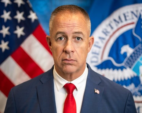 a man in a suit looks straight ahead, an american flag and flag for the department of homeland security behind him