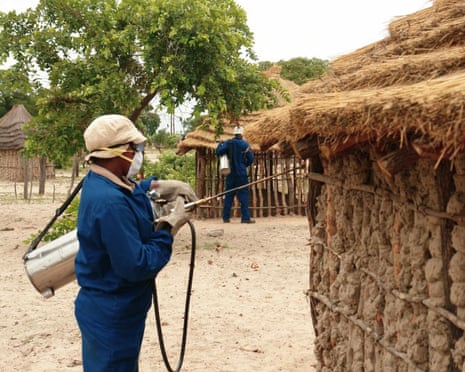 A person wearing protective gear sprays a small building with a thatched roof