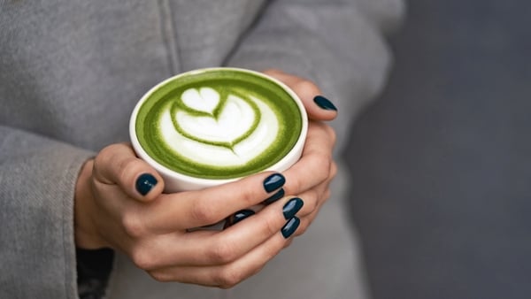 Hands of woman in gray coat hold a ceramic cup with matcha green tea latte with foam art. Backdrop for your design with copy space. Close-up photography from above